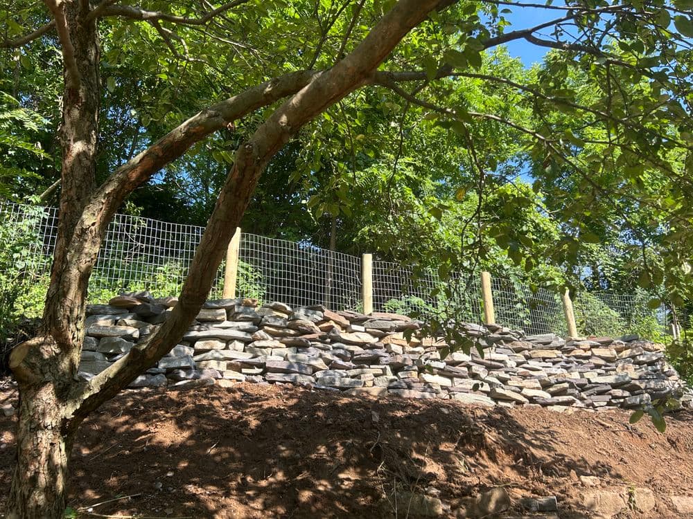 Stone retaining wall with fencing, surrounded by lush greenery under a clear blue sky.
