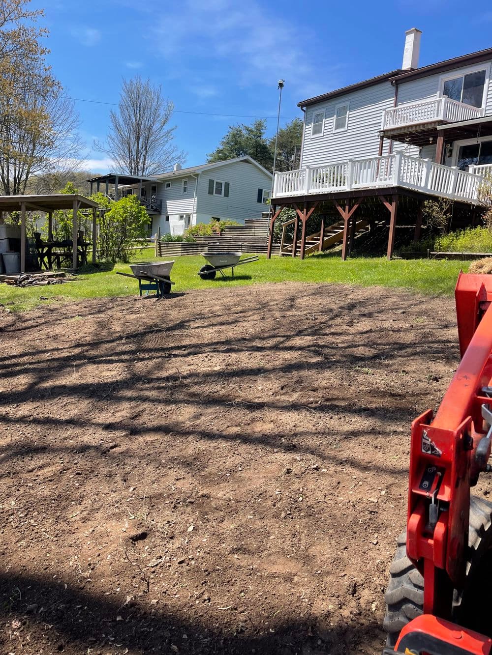 Landscaped backyard with a tractor, wheelbarrow, and two houses in the background on a sunny day.