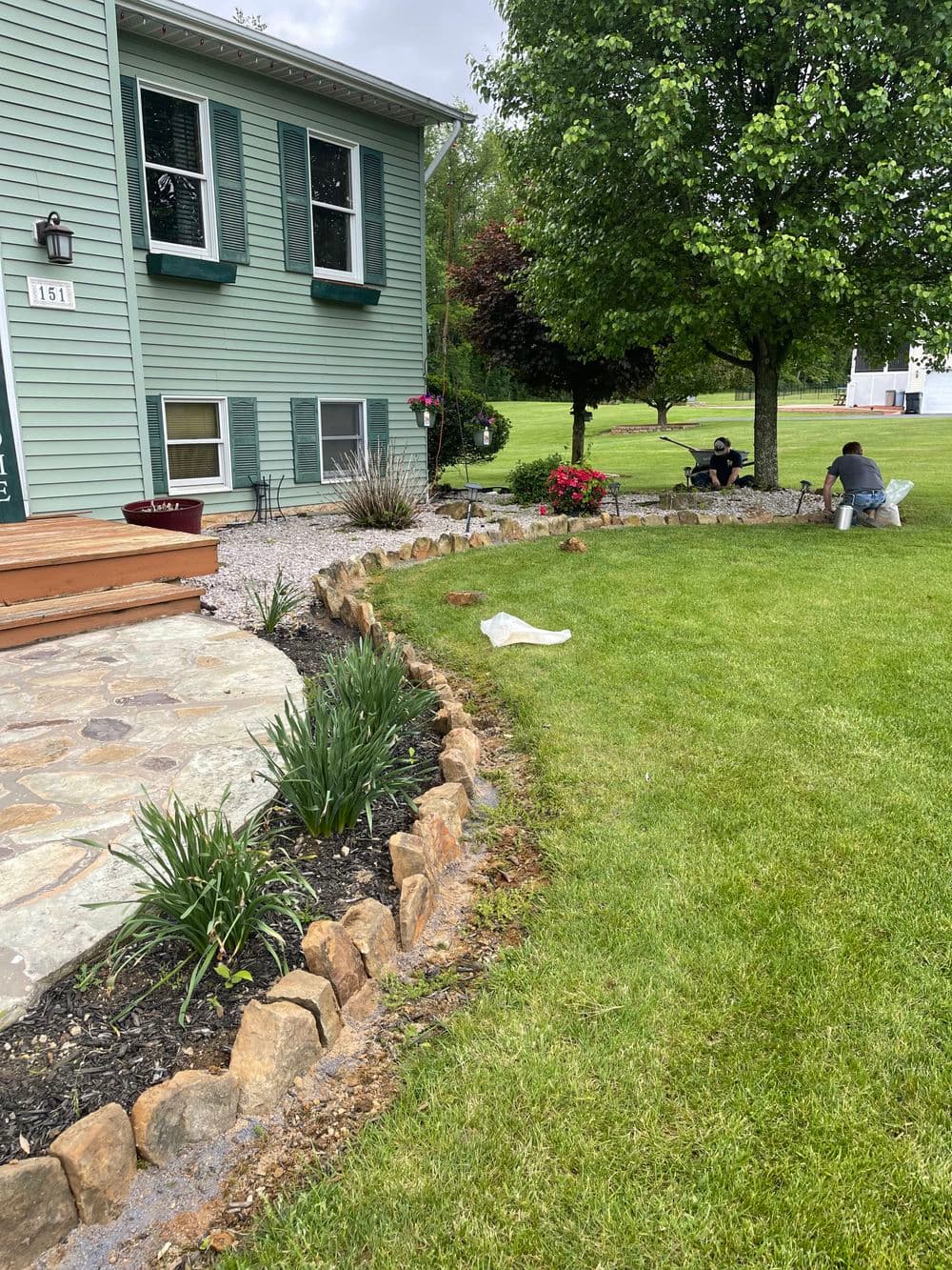 Lush green lawn with stone landscaping and workers tending to garden near a blue house.