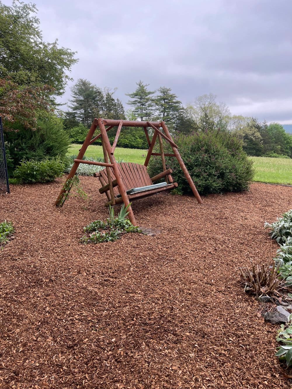 Wooden swing seat in a landscaped yard with mulch and greenery under a cloudy sky.